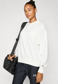 Pullove bianco con colletto e logo sul petto, abbinato a pantaloni sportivi scuri e una borsa a tracolla nera texture.