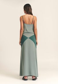 Ærmeløs, rygfri maxikjole i lys grøn, med mørkere grønne geometriske paneler, diskret glans og en lige silhuet.