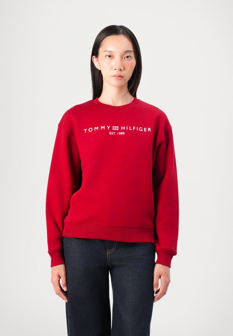 Červená mikina s kulatým výstřihem a žebrovanými manžetami, na středu s bílým logem "TOMMY HILFIGER EST. 1985". Kombinováno s tmavými džíny.