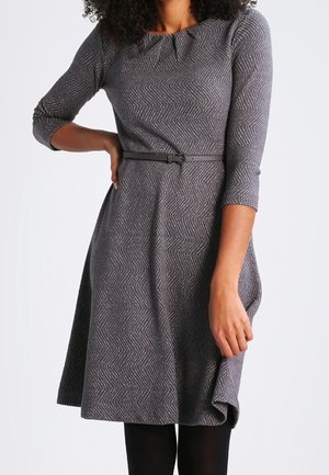 Femme portant une robe grise ajustée avec un motif chevron subtil, des manches trois-quarts et une fine ceinture noire à la taille.