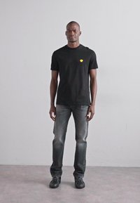 Μαύρο κοντομάνικο μπλουζάκι με μικρό κίτρινο λογότυπο, συνδυασμένο με ξεθωριασμένα μπλε τζιν και μαύρα παπούτσια. Απλό φόντο και neutra φωτισμός.