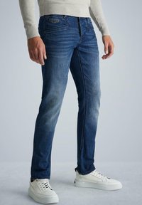 Vaqueros de mezclilla azul con ajuste slim, desgaste ligero y detalles sutiles de desgaste, combinados con zapatillas blancas y un suéter de punto de color claro.