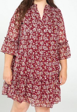 Woman wearing a loose, knee-length red dress with white floral print and three-quarter ruffled sleeves, standing against a plain background.