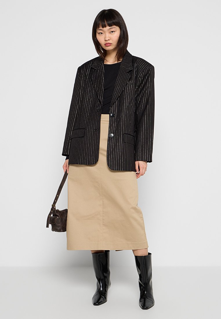 Czarna marynarka w paski założona na czarną bluzkę, zestawiona z beżową spódnicą midi i czarnymi botkami za kolano. Modelka trzyma małą torebkę.