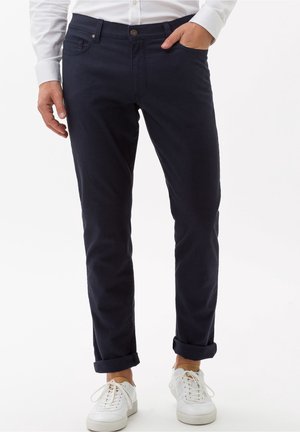 Pantalons en coton bleu marine à coupe slim, avec fermeture à bouton et revers roulés. Associés à des baskets blanches, offrant un look décontracté.