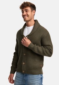 Oliwkowy sweter z dzianiny z kołnierzem szalowym, z drewnianymi guzikami i teksturowanym wzorem. Noszony rozpięty na białym T-shircie.