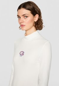 Hvid langærmet skjorte med høj krave, prydet med et lille broderet "G" logo i rød og blå. Glat, tætsiddende tekstur.