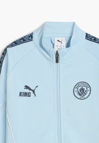 Ljusblå zip-up-jacka med marinblå detaljer, prydd med Puma-logotyp, texten "KING" och Manchester City-emblem. Slät tygstruktur.