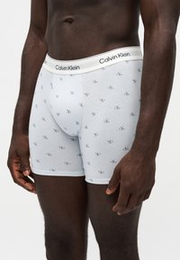 Homme portant un boxer blanc Calvin Klein avec un petit motif répétitif du logo "CK" et une ceinture de marque.