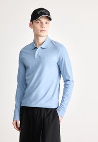 Šviesiai mėlyna ilgomis rankovėmis polo marškinėliai su apykakle ir maža logotipo žyme. Dėvimi su juodais šortais. Modelis pozuoja priešais paprastą foną.