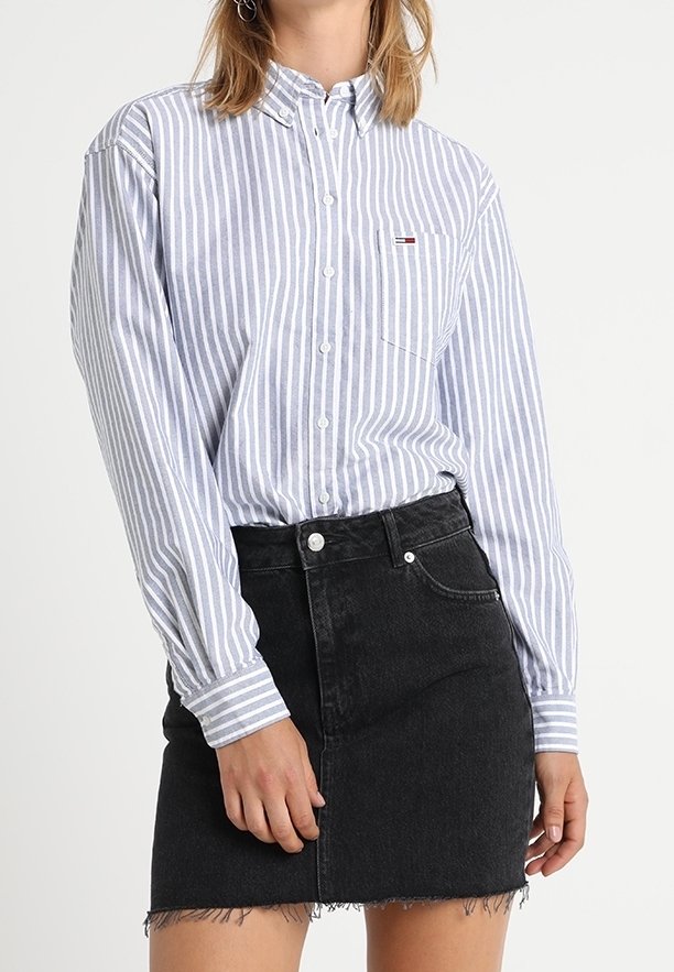 Camicia a maniche lunghe a righe blu chiaro e bianche con taschino sul petto, abbinata a una minigonna di denim nera con orli sfrangiati.