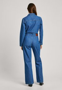 Denim kombinezon v temno modri barvi, z dolgimi rokavi, oprijetim pasom, širokimi hlačami in usnjenim znakom na hrbtu. Gladka tekstura.