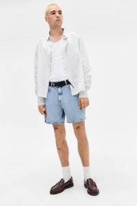 Ungdom med kort blondt hår som har på seg en hvit singlet, stripete skjorte med knapper, lyse denimshorts, hvite sokker og brune loafers, står.