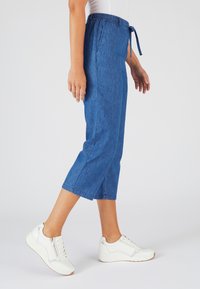 Pantalones capri de mezclilla en un tono azul medio, con cintura ajustable y bolsillos laterales. Combinados con zapatillas blancas, presentan un look casual.