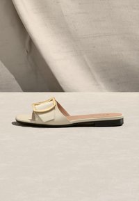 Sandale plate beige à bride large, grande boucle rectangulaire dorée, et semelle noire sur un fond en tissu neutre.