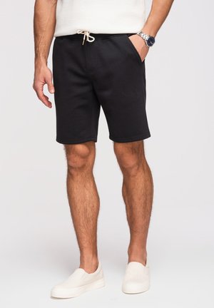 Hombre llevando pantalones cortos negros con cordón, zapatos blancos sin cordones, reloj plateado y camiseta sin mangas color crema, de pie con una mano en el bolsillo.