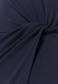 Námořnicky modrá látka s hladkým povrchem, která má uzlový design v centru, vytvářející nařasené záhyby a jemné drapérie.