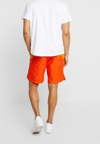 Pánské oranžové plavky s lehkou, lesklou texturou, vybavené postranními kapsami a elastickým pasem. Kombinovány s bílým tričkem a teniskami.