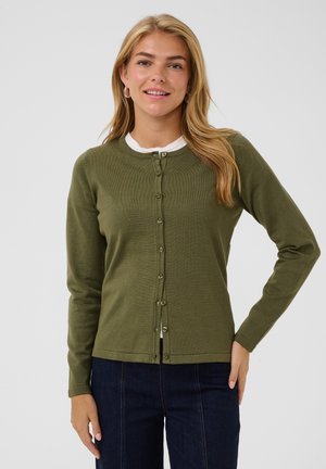 Giovane donna con lunghi capelli biondi che indossa un cardigan verde oliva con bottoni sopra una camicia bianca e jeans blu scuro, in piedi davanti a uno sfondo semplice.