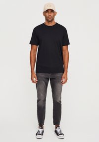 Černé tričko s krátkým rukávem, šedé slim-fit džíny a černo-bílé tenisky. Béžová kšiltovka s logem, stojící na jednobarevném pozadí.