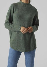 Femme portant un pull vert ample à col rond côtelé et un jean bleu, debout les mains croisées à la taille devant un fond uni.