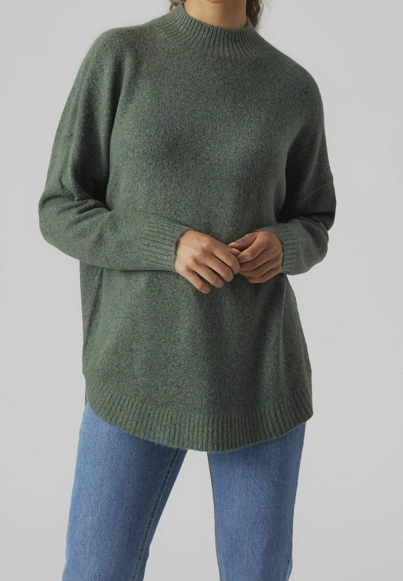 Femme portant un pull vert ample à col rond côtelé et un jean bleu, debout les mains croisées à la taille devant un fond uni.