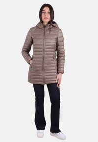 Giacca imbottita effetto cappotto fino al ginocchio di colore marrone chiaro con cappuccio, caratterizzata da cuciture orizzontali, cerniera frontale e tasche laterali. Abbinata a jeans scuri e scarpe bianche.