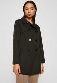 Impermeabile nero doppiopetto con colletto, grandi bottoni e tasche laterali. Realizzato in tessuto liscio, presenta cuciture sartoriali e una vestibilità slim.