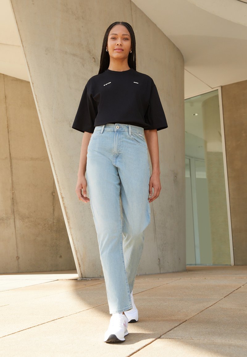 T-shirt oversized preto com letras brancas, jeans de cintura alta azul-claro e ténis brancos; tecido suave, ajuste descontraído e corte reto.