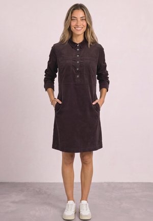 Femme souriante portant une robe brun foncé à manches longues, boutonnée avec col, et des baskets blanches, debout les mains dans les poches.
