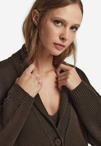 Mörkbrun cardigan med en strukturerad stickad design, med djup V-hals, knappstängning och ribbade ärmslut. Bärs över en matchande topp.