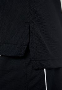 Černé sportovní oblečení s texturovanou síťovinou a detailem rozparku na spodním okraji. Na spodním lemu jsou viditelné kontrastní bílé akcenty.