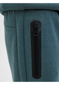 Teal jogger kalhoty vyrobené z měkké textilie s bočním zipovým kapsou, která má černý zip a texturovaný povrch.