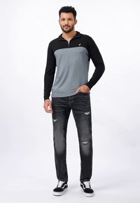 Ilgos rankovės pilkos ir juodos spalvos megztinis su užtrauktuku ir aukštu apykakliu, derinamas su pažeistais juodais džinsais ir juodais sportbačiais.