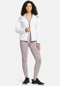Biela bunda na zips s čiernymi akcentmi, v kombinácii s svetloružovými legínami a čiernymi športovými topánkami. Modelka má kučeravé vlasy a stojí vzpriamene.