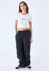 T-shirt blanc court avec le texte noir "Project x Paris" associé à un pantalon cargo noir ample et des baskets noires avec des accents blancs.