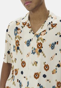 Korte ærmer skjorte i let stof med en cremefarvet bund og farverige blomster mønstre i blå, brun, grøn og fersken. Klassisk krave og knapper.