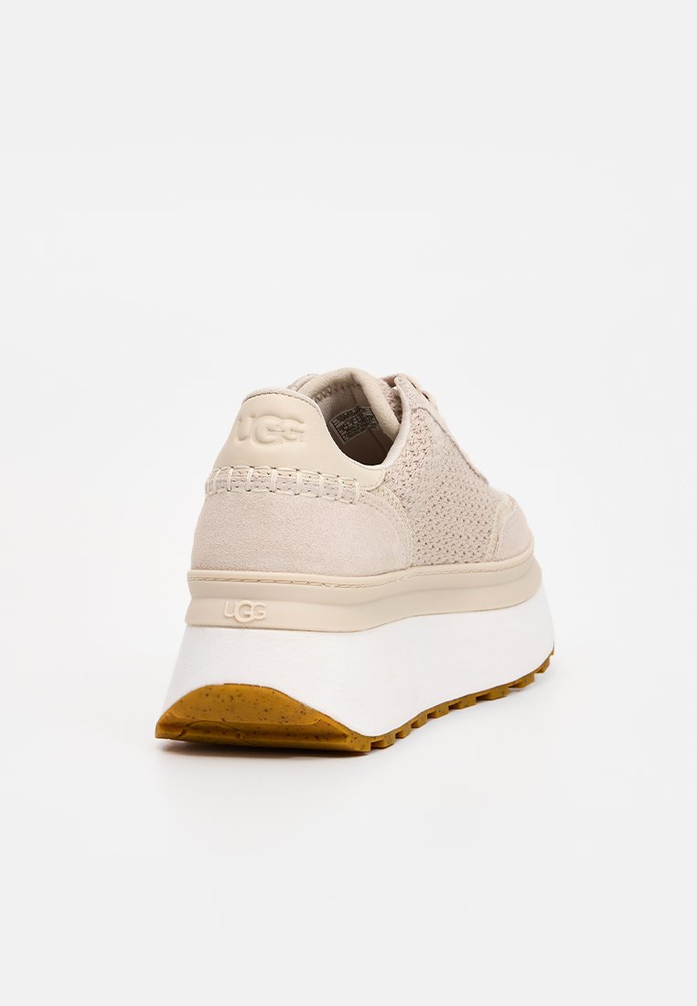 Sneaker mit einem hellbeigen strukturierten Obermaterial, einer dicken weißen Zwischensohle und einer braunen Gummisohle. UGG-Logo auf der Ferse geprägt.