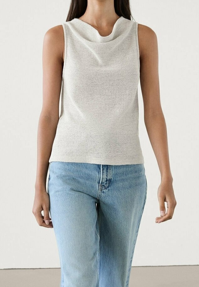 Woman wearing a sleeveless white knit top with a draped neckline and light blue high-waisted jeans against a plain background.