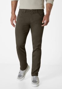 Men's dark olive green pants made of sturdy fabric, featuring a slim fit, five-pocket design, and a classic button fly. Paired with grey sneakers.