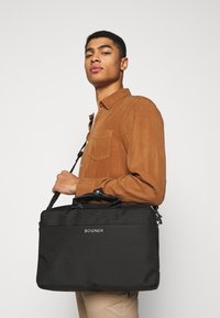 Sort laptop taske med tekstureret overflade og dobbelte håndtag; har en skulderrem og et prægede logo på forsiden.