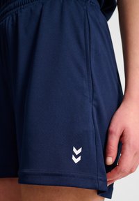 Marina idrottsshorts gjorda av slät väv; har en elastisk midja och en vit logotyp med två chevronformer på den nedre sidan.