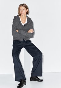 Grauer Strickcardigan mit Kapuze und Knöpfen, kombiniert mit schwarzen gestreiften Hosen und glänzenden schwarzen Slippern, vor einem weißen Hintergrund.