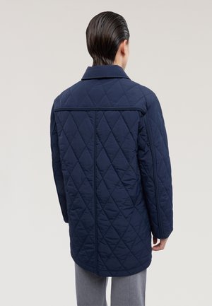 Marineblå quiltet jakke med krave, der har et diamantmønster og en midterlang pasform. Stoffet virker glat og let.