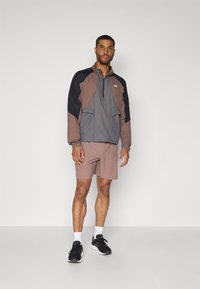 Sportjacka med halvdragkedja, grå och brun färgblockdesign. Kombinerad med ljusbruna shorts och svarta träningsskor.