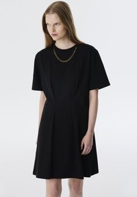 Schwarz kurzes Kleid mit kurzen Ärmeln, tailliertem Oberteil und schwingendem Rock. Verfügt über einen runden Ausschnitt und einen goldenen Kettenakzent am Kragen.