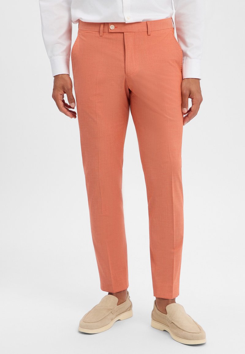 Mann trägt schmale Korallenhose, weißes Hemd und beige Wildleder-Loafer, steht vor hellem Hintergrund.