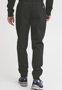 Pantalons de survêtement noirs avec un design fuselé, une taille ajustable et une petite étiquette de logo sur la poche arrière, associés à des sneakers de sport.