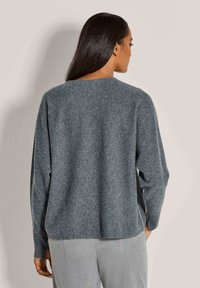 Frau mit langen dunklen Haaren, die einen lockeren grauen Pullover und helle graue Hosen trägt, von hinten gegen einen schlichten Hintergrund betrachtet.