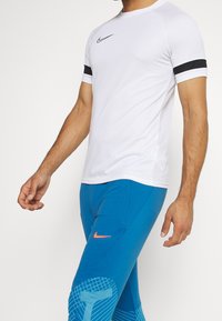 Witte sportieve T-shirt met zwart-wit gestreepte mouwen, vergezeld van blauwe leggings met een gepatterneerd kniegedeelte en een multicolore logo.